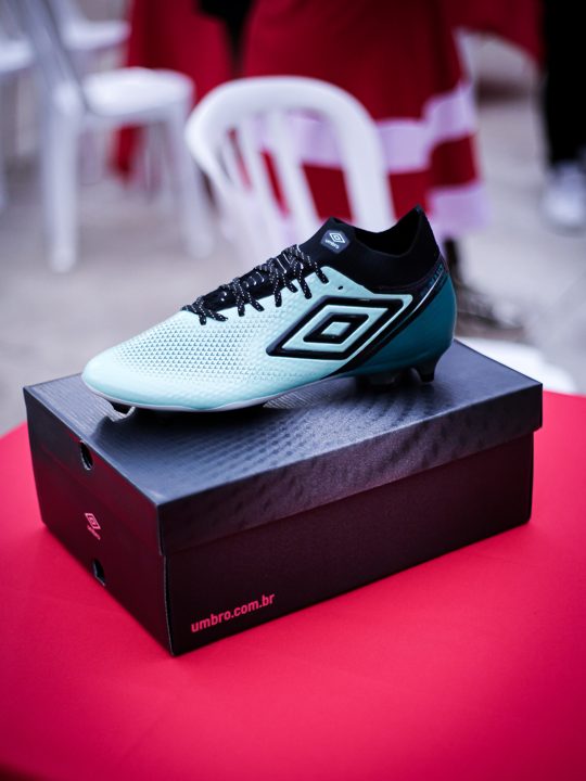 Chuteira verde sobre a caixa Umbro em uma cadeira vermelha no espaço do encontro. Registro do Churrasco Fair Play, no acervo da SCP 2026.