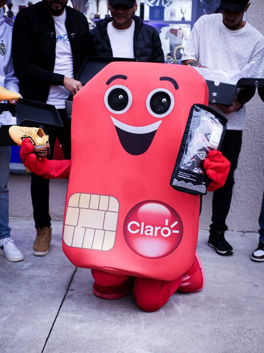 Mascote da Claro apresenta uma chuteira dourada Umbro, com participantes e embalagens ao fundo. Registro do Churrasco Fair Play, no acervo da SCP 2026.
