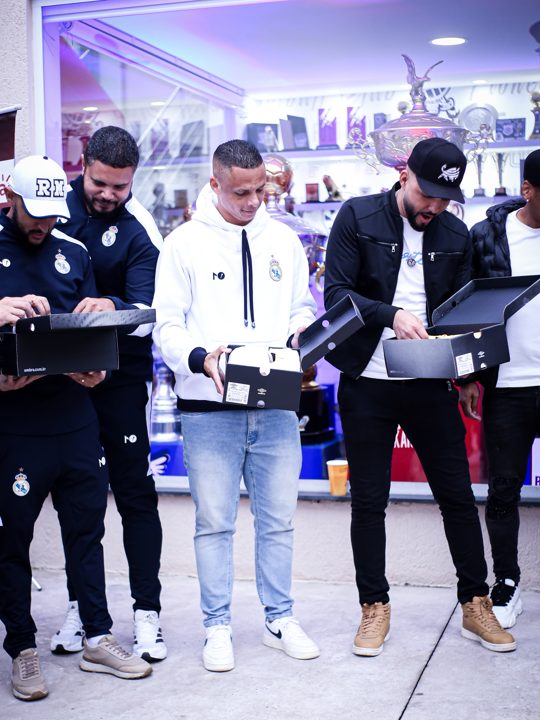 Participantes abrem caixas com o duplo diamante Umbro diante do espaço de troféus. Registro do Churrasco Fair Play, no acervo da SCP 2026.