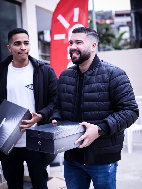 Dois participantes posam segurando caixas fechadas da Umbro, com o duplo diamante visível. Registro do Churrasco Fair Play, no acervo da SCP 2026.