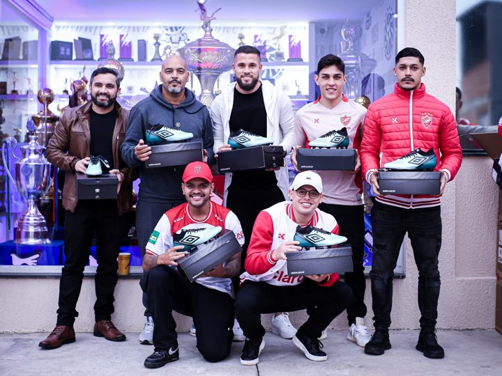 Grupo de participantes apresenta chuteiras verdes Umbro e caixas da marca diante dos troféus.