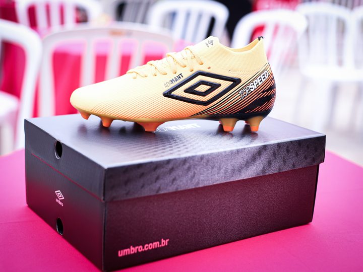 Detalhe lateral de uma chuteira Umbro dourada sobre sua caixa, com o duplo diamante em destaque.