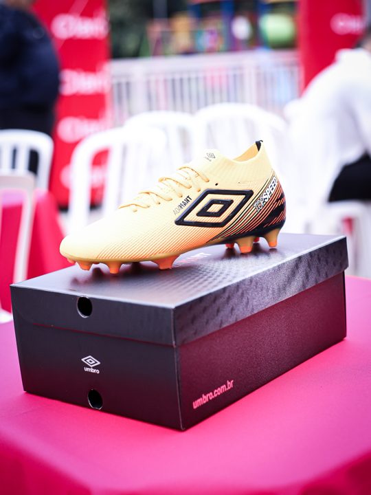 Composição vertical de uma chuteira Umbro dourada apoiada na embalagem preta, com cadeiras do encontro ao fundo.
