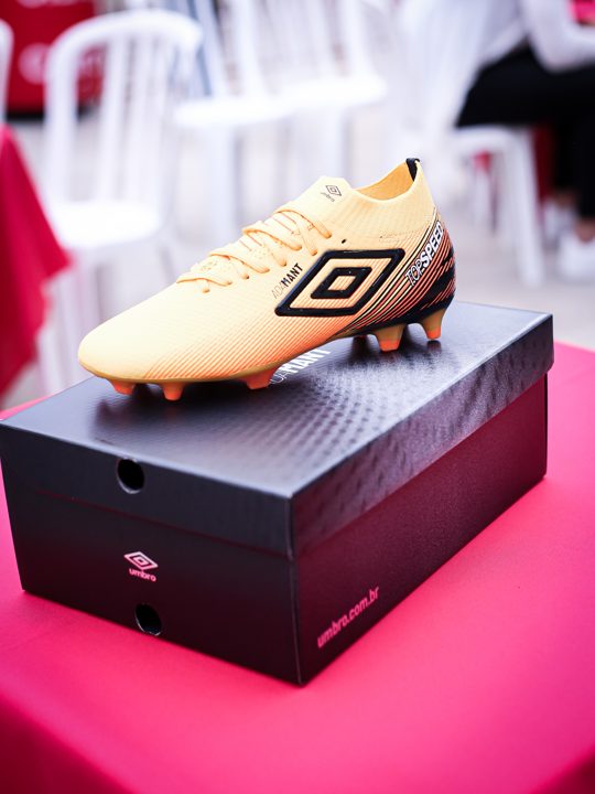 Chuteira dourada Umbro e caixa fotografadas sobre uma superfície vermelha durante o encontro.