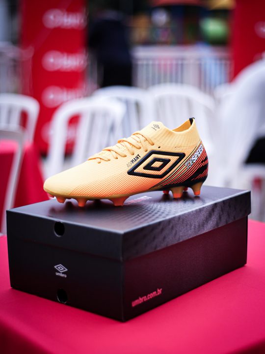 Detalhe do produto Umbro sobre a caixa preta, com a marca visível na lateral da chuteira dourada.