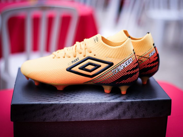Par de chuteiras douradas Umbro sobre a caixa, com o duplo diamante e o grafismo lateral dos produtos visíveis.