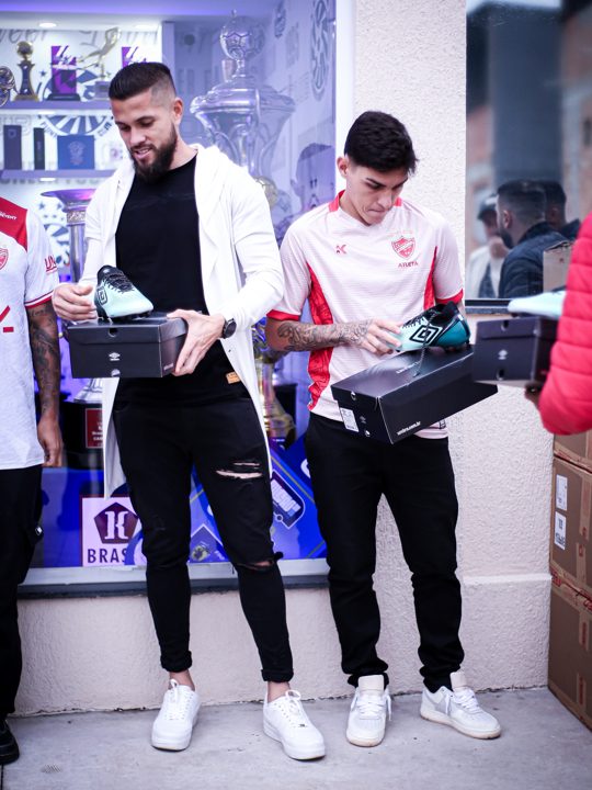 Dois participantes observam chuteiras verdes Umbro e suas embalagens, diante da sala de troféus.