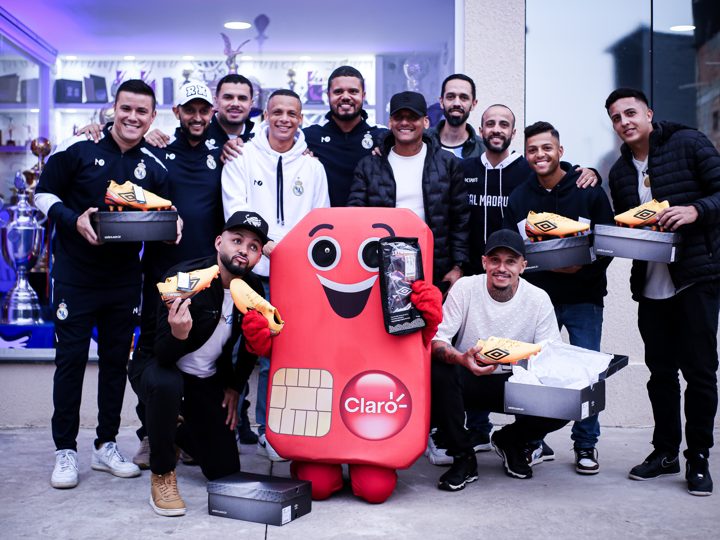 Registro coletivo de participantes com chuteiras douradas Umbro e suas embalagens, ao lado da mascote da Claro.