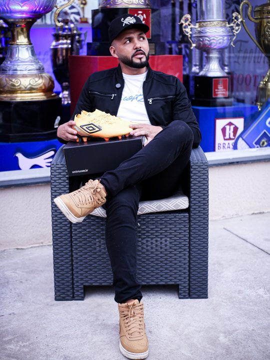 Participante sentado segura uma caixa com a chuteira Umbro dourada apoiada sobre a tampa, diante de troféus.