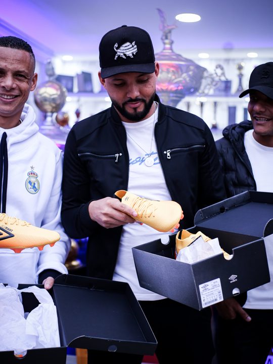 Participante de jaqueta preta observa uma chuteira dourada ao lado de caixas da Umbro, no encontro com participantes da SCP.
