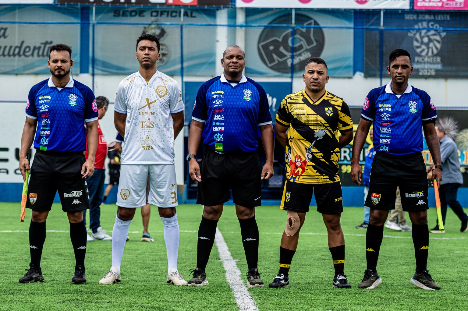 Trio de arbitragem com aplicação do logotipo Umbro nas camisas, ao lado de atletas. O registro comprova a aplicação visível na arbitragem; o enquadramento na obrigação de camisas da organização e a fase precisam ser validados.
