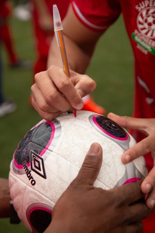 Detalhe de uma bola Umbro sendo assinada por uma pessoa com camisa da Super Copa Pioneer. O logotipo da marca aparece em primeiro plano.