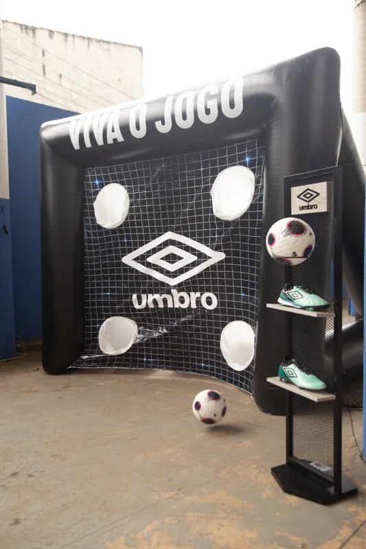 Estrutura inflável de alvo com a frase “Viva o Jogo” e identidade Umbro, ao lado de um expositor com bola e chuteiras da marca.