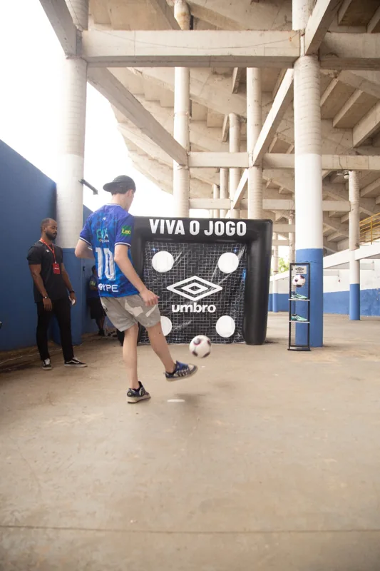 Participante de camisa azul realiza um chute em direção ao alvo “Viva o Jogo”, identificado com a marca Umbro. Um expositor de produtos aparece ao lado.