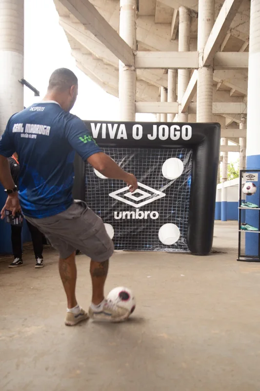 Participante com camisa identificada como Real Madruga chuta uma bola diante do alvo Umbro “Viva o Jogo”.
