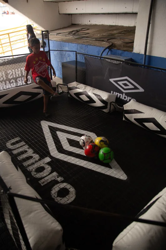 Criança participa de uma brincadeira com bolas em espaço com pequenos gols e identidade Umbro aplicada ao piso, às proteções e ao painel lateral.