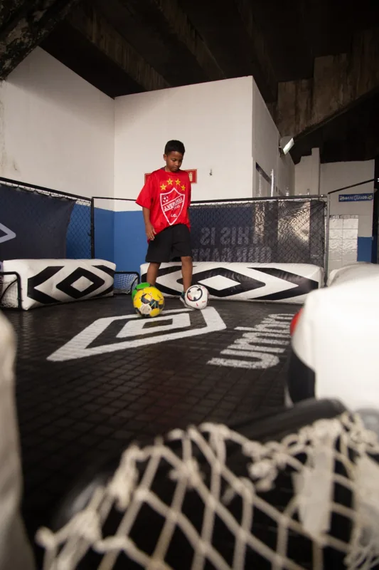 Criança com camisa vermelha brinca com bolas no espaço identificado pela marca Umbro, com um pequeno gol em primeiro plano.