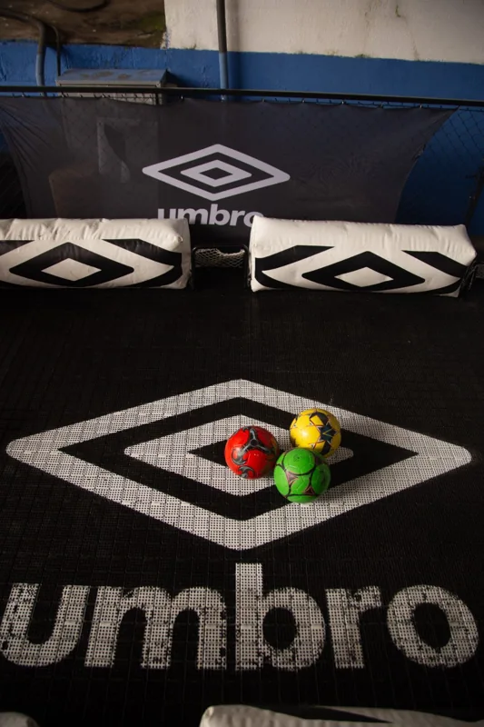 Bolas coloridas sobre piso com o duplo diamante e o nome Umbro, diante de proteções e painel também identificados com a marca.