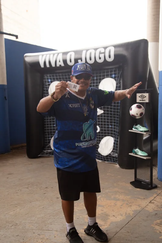 Participante posa diante do alvo “Viva o Jogo” e do expositor Umbro, segurando um cartão com QR code.