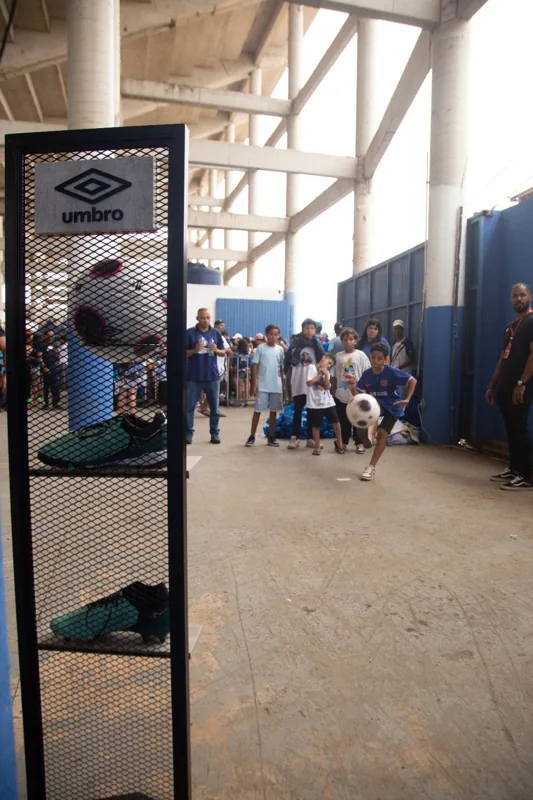 Criança chuta uma bola enquanto outras pessoas acompanham a atividade. Em primeiro plano, um expositor apresenta a marca Umbro, uma bola e chuteiras.