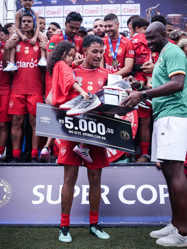 Jogador de uniforme vermelho, acompanhado de uma criança, recebe produtos Umbro diante da equipe com medalhas. A placa exibe “Vale compras de R$ 5.000,00 no site umbro.com.br”.