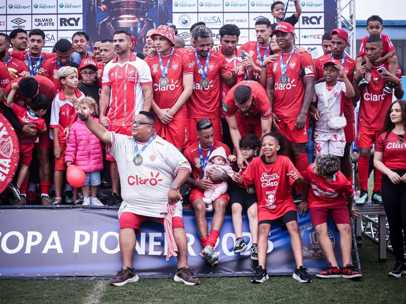 Equipe de uniformes vermelhos e acompanhantes reunidos no pódio, com medalhas e painel de patrocinadores ao fundo. A marca Umbro aparece no painel.