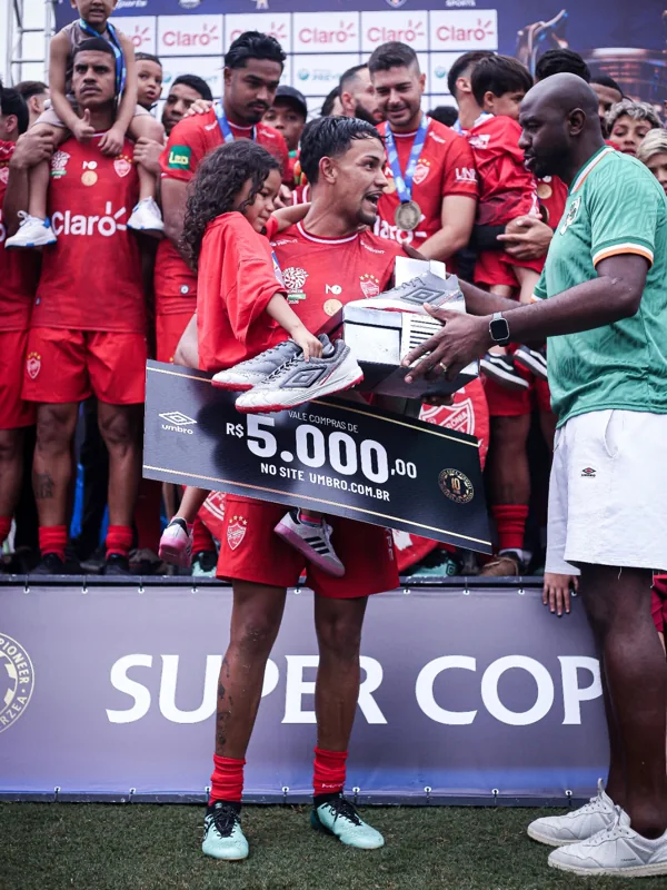 Outro enquadramento da entrega de produtos Umbro ao jogador de uniforme vermelho, com a placa de vale-compras de R$ 5.000,00 em destaque.