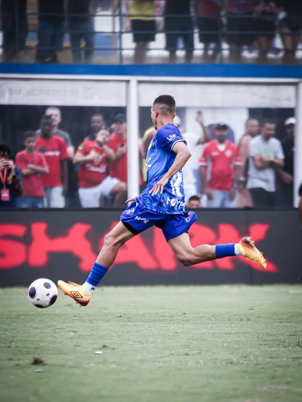 Jogador de uniforme azul executa um chute com chuteiras douradas Umbro. A bola branca com detalhes pretos e rosas aparece à frente do pé.