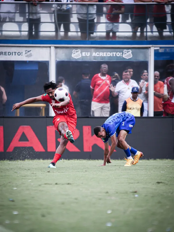 Jogador de uniforme vermelho disputa um lance com jogador de azul, que utiliza chuteiras douradas com o duplo diamante Umbro visível. A bola aparece em movimento.