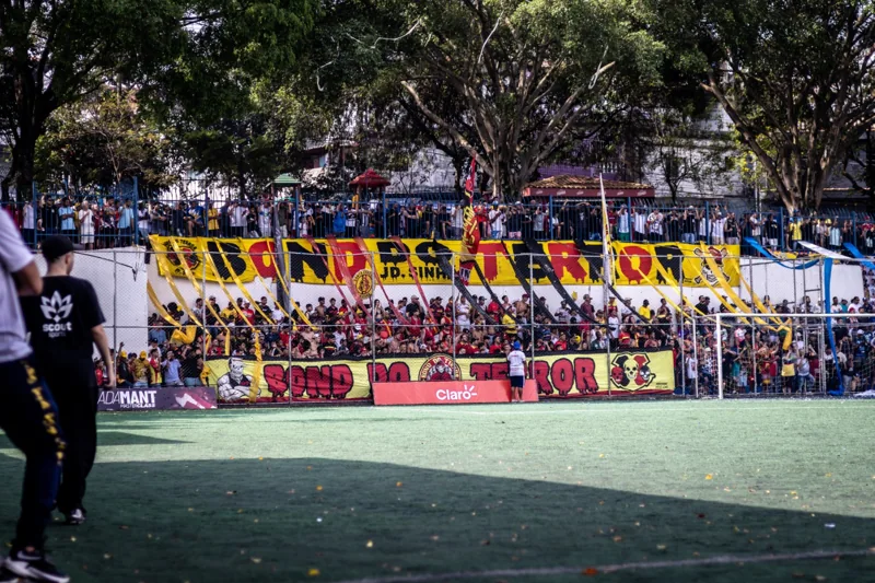 Torcida junto ao alambrado e às faixas da arquibancada. À beira do campo, uma placa exibe “ADAMANT MASTERCLASS”, linha de chuteiras da Umbro identificada em fonte oficial.