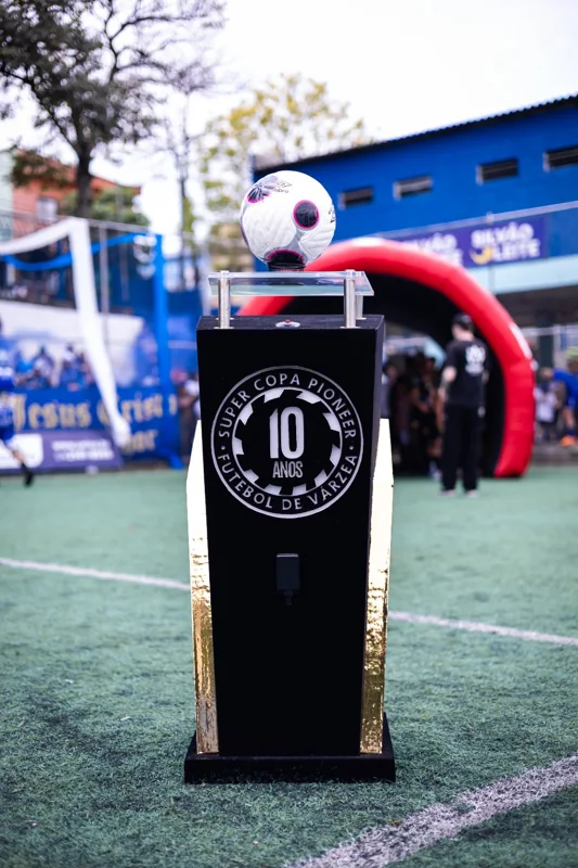 Bola branca Umbro com detalhes pretos e rosas sobre um pedestal identificado com os 10 anos da Super Copa Pioneer, instalado no campo.