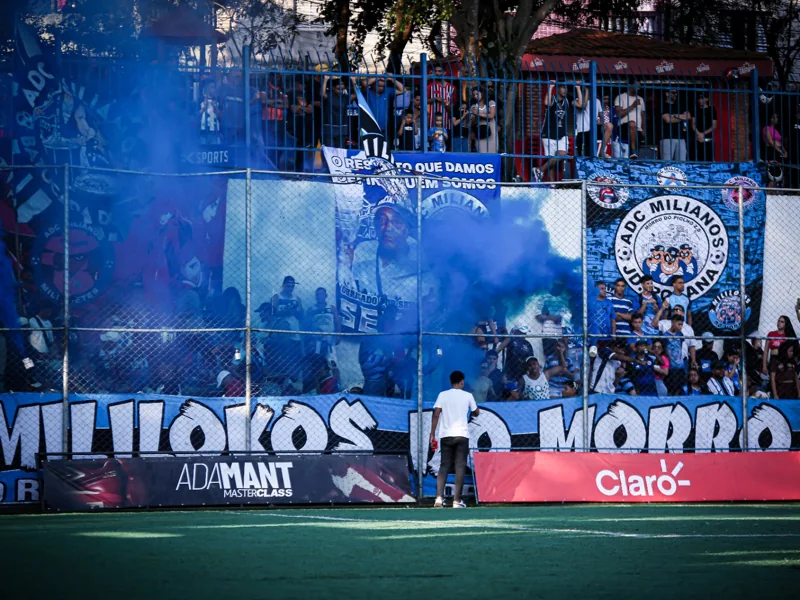Torcida com faixas azuis e fumaça na arquibancada. Diante do alambrado, aparece uma placa “ADAMANT MASTERCLASS” com imagem de calçado.