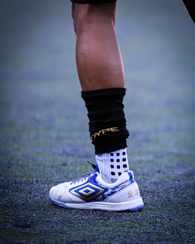 Detalhe de calçado branco, azul e dourado em uso no gramado, com o duplo diamante Umbro em destaque na lateral.