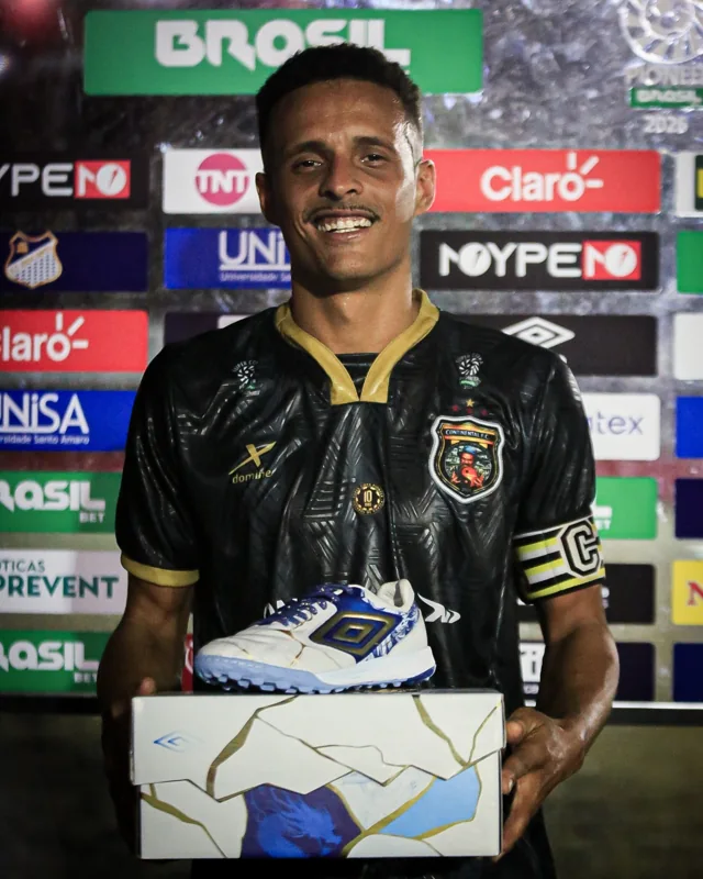 Atleta com camisa identificada como Continental FC segura uma caixa com calçado branco, azul e dourado Umbro. Um duplo diamante também aparece no painel de patrocinadores ao fundo.