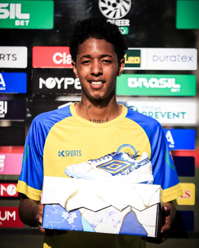 Atleta com camisa amarela e azul segura caixa e calçado Umbro branco, azul e dourado diante de um painel de patrocinadores.