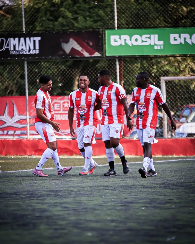 Quatro jogadores de uniformes listrados em vermelho e branco caminham juntos no campo. No alambrado ao fundo aparece uma placa “ADAMANT MASTERCLASS”, parcialmente enquadrada.