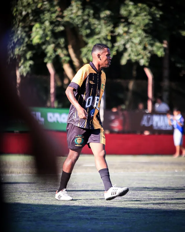 Jogador de uniforme preto e dourado em movimento no campo, com placas publicitárias desfocadas ao fundo. Registro de contexto geral da competição.