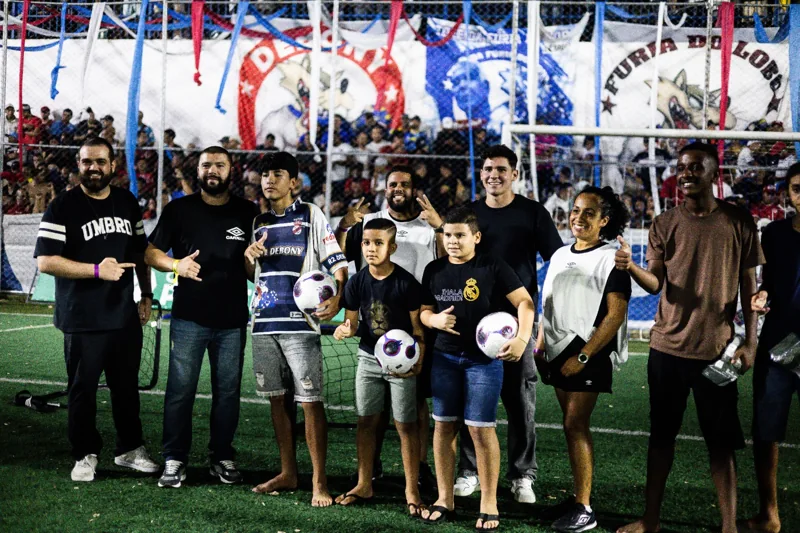 Participantes posam em grupo no campo junto a pequenos gols. Três jovens seguram bolas; o nome Umbro e o duplo diamante aparecem em peças de roupa de participantes.