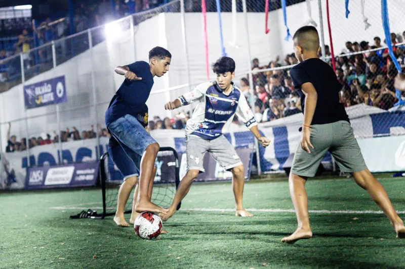 Três jovens disputam uma bola junto a um pequeno gol no campo, com público nas arquibancadas ao fundo. Registro de contexto geral da competição.