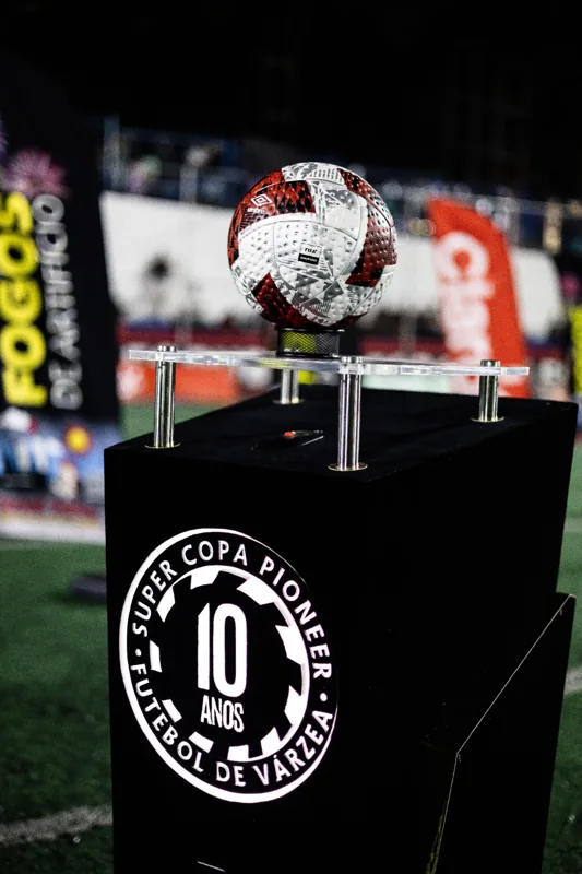 Bola branca, vermelha e preta Umbro em exposição sobre o pedestal dos 10 anos da Super Copa Pioneer, em registro noturno.