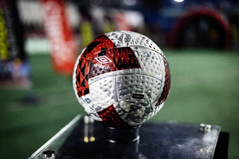 Enquadramento horizontal da bola Umbro sobre o pedestal SCP 10 anos. O nome da marca e o duplo diamante aparecem no produto.
