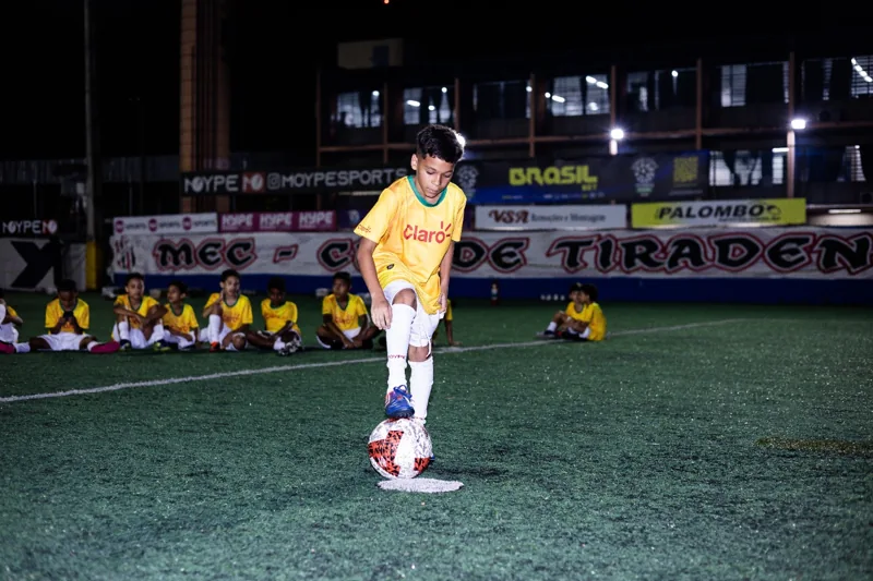Criança de uniforme amarelo apoia o pé sobre uma bola branca, vermelha e preta; outras crianças acompanham a atividade sentadas no gramado. Registro de contexto geral da competição.