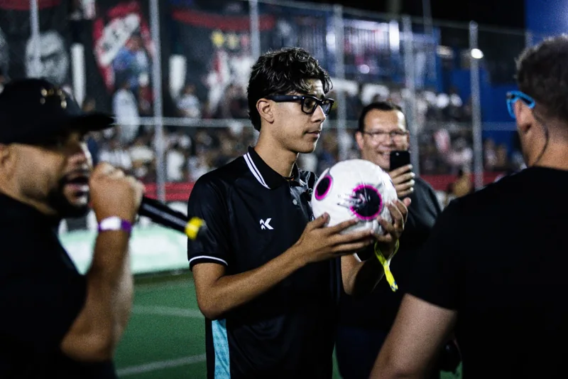 Participante de camisa polo preta segura uma bola embalada, branca com detalhes escuros e rosas, durante uma interação no campo. Registro de contexto geral da competição.