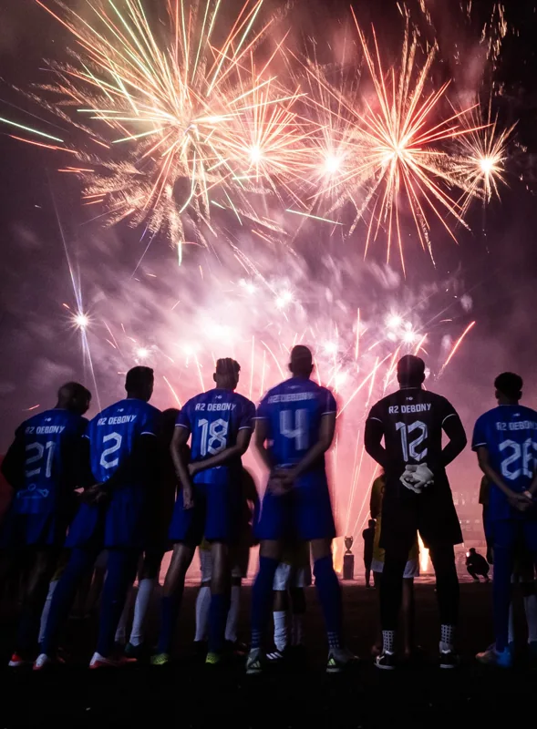 Jogadores de uniformes azuis acompanham uma queima de fogos junto ao campo durante um registro noturno da competição. Registro de contexto geral da competição.
