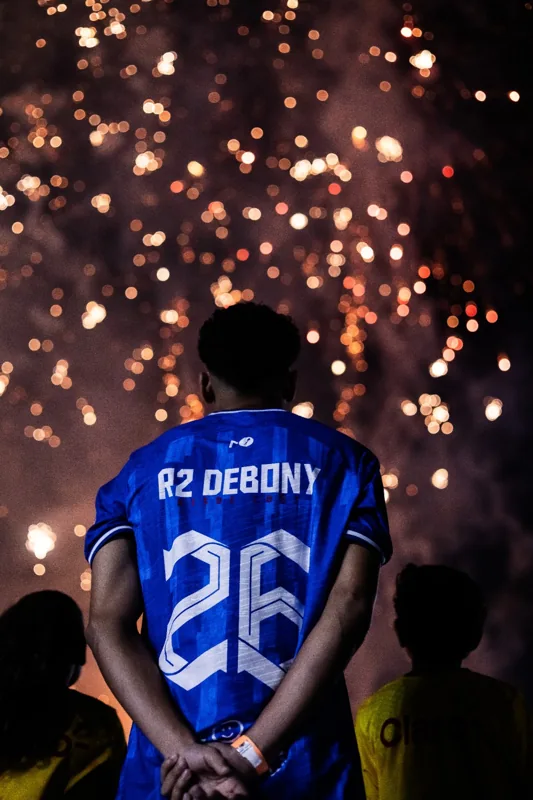 Jogador com a camisa azul número 26 aparece de costas, com os fogos ao fundo. Registro de contexto geral da competição.