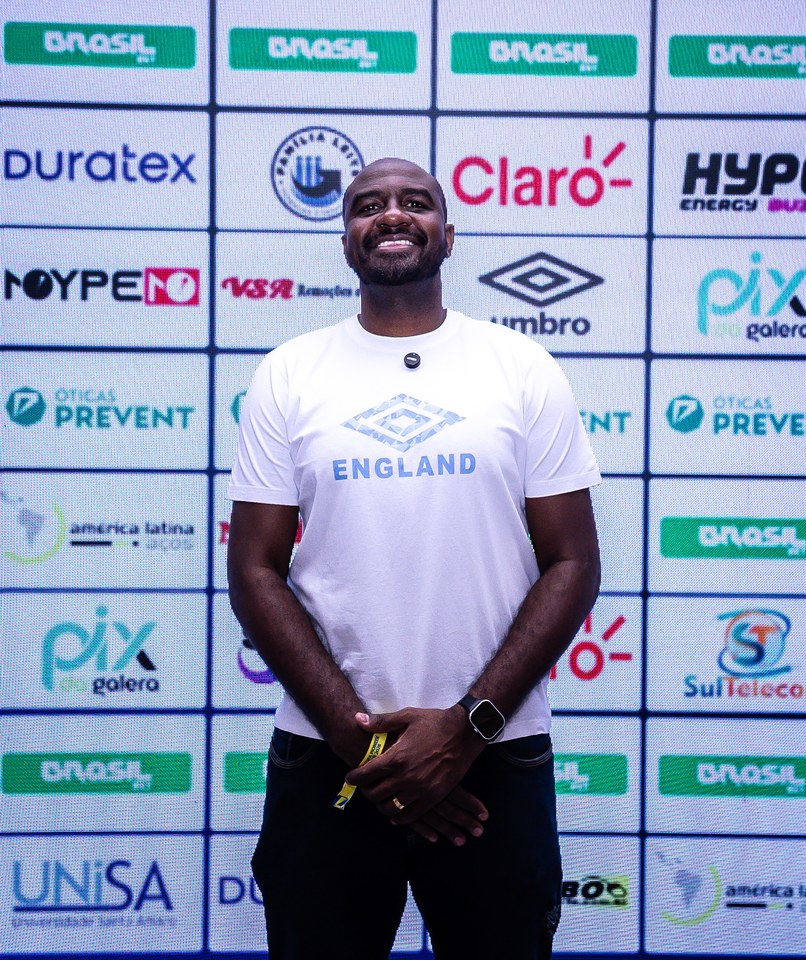 Participante com camiseta Umbro posa diante do painel de patrocinadores do Congresso Técnico da SCP 2026. A marca também aparece no painel ao fundo.