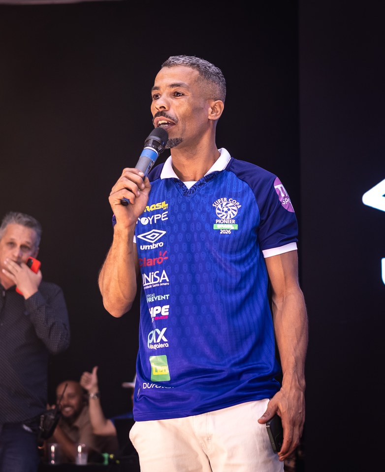 Participante ao microfone com camisa azul da SCP 2026. O logotipo Umbro aparece na frente da peça, entre os patrocinadores. A foto registra a aplicação; o enquadramento na entrega de camisas da organização permanece a validar.