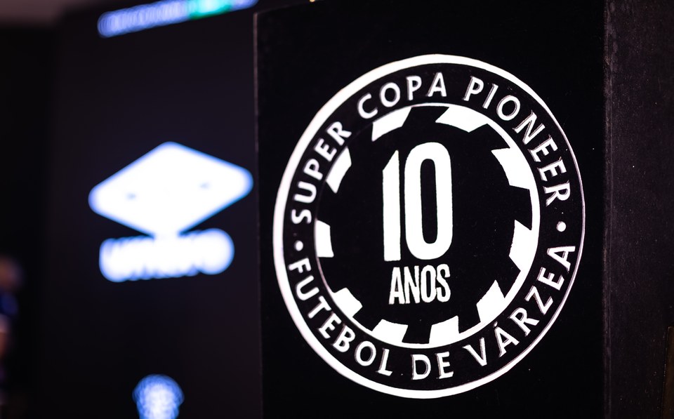 Símbolo dos 10 anos da Super Copa Pioneer em foco, com a marca Umbro desfocada no painel ao fundo. Fotografia editorial complementar do Congresso Técnico.