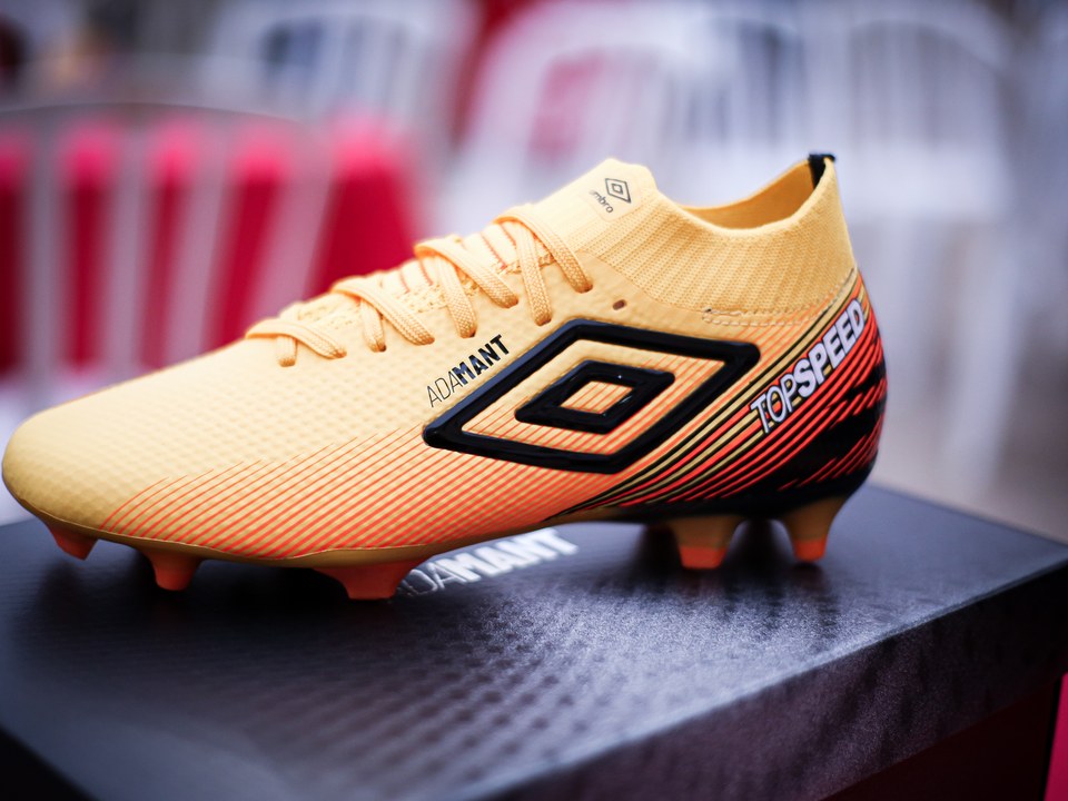 Chuteira Umbro Adamant Top Speed amarela sobre sua caixa, fotografada no encontro do acervo “Churrasco Claro + Chuteira Umbro” da SCP 2026. O duplo diamante aparece na lateral e na língua do produto.