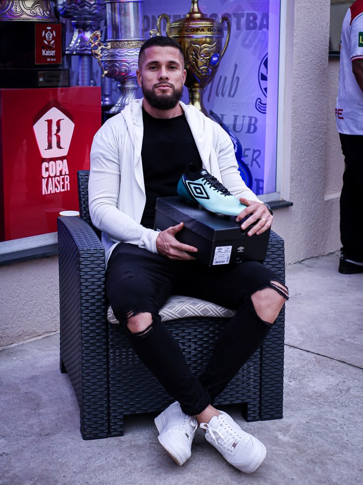 Participante apresenta uma chuteira azul-clara e a embalagem Umbro diante de uma vitrine de troféus. Fotografia do acervo “Churrasco Claro + Chuteira Umbro” da SCP 2026; identificação do participante e critério de entrega a confirmar.